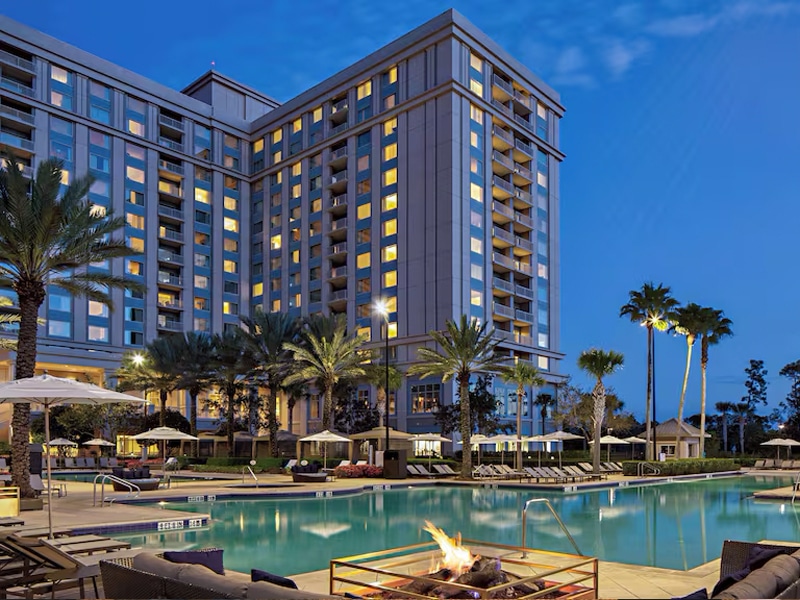 Evening view of the Waldorf Astoria Orlando with its lit windows, poolside firepit, and palm trees surrounding a calm resort-style swimming pool.