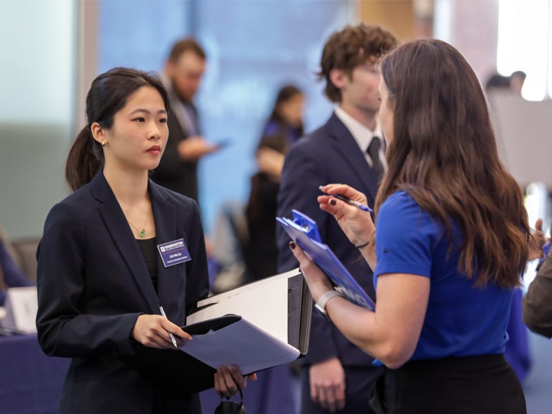 Student in business attire speaks with a recruiter during a career fair, holding a notepad and resume folder.