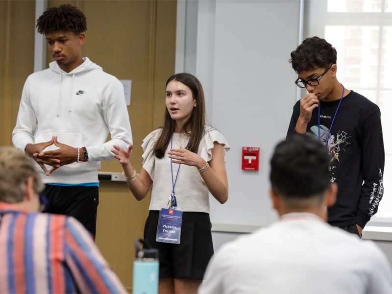 Three students stand and present to a classroom audience, with one student speaking while the others listen attentively.