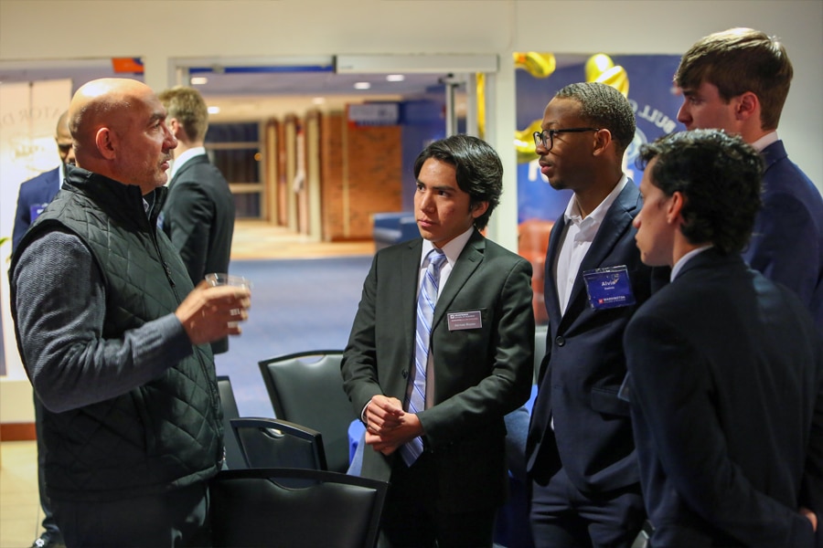 A group of undergraduate students in business attire attentively listen to an industry advisor during a networking session at the Future Advisor Conference.