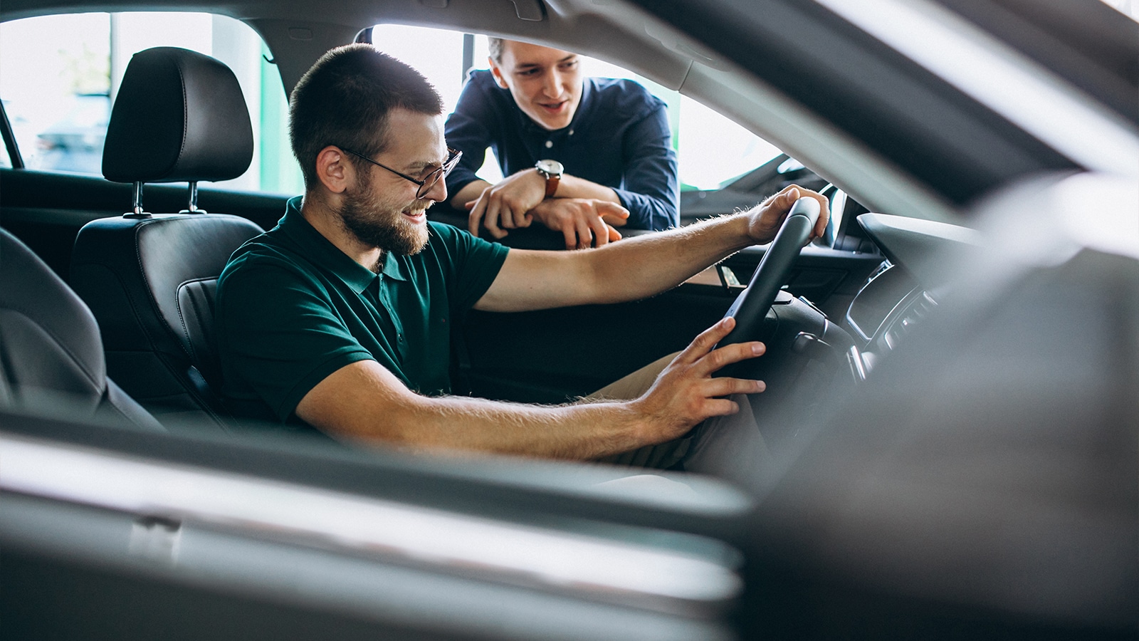 A smiling man sits in the driver’s seat of a new car, testing the steering wheel while another man, possibly a salesperson or friend, stands outside the car offering guidance.