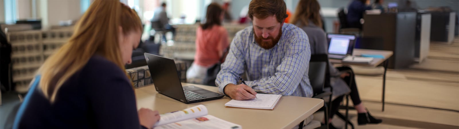 Two graduate students study at a table with laptops and notebooks in a quiet academic setting.