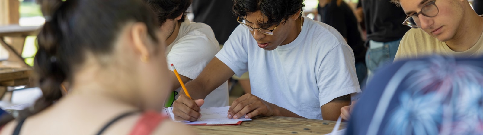 Students seated at a picnic table writing in notebooks during an outdoor workshop.