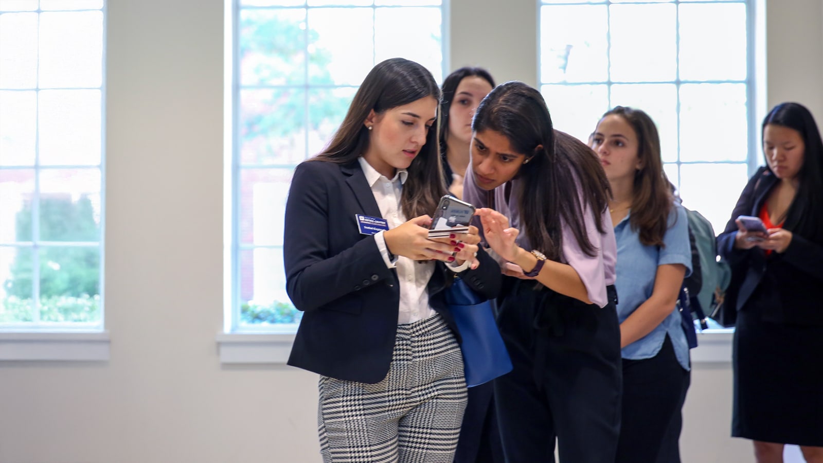 Two students review information on a mobile phone while others wait in line behind them in a brightly lit room.