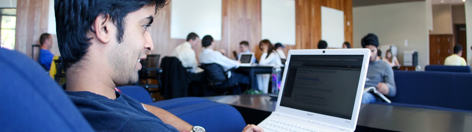 A student works on a laptop in a modern lounge area at Hough Hall, with other students studying and collaborating in the background.