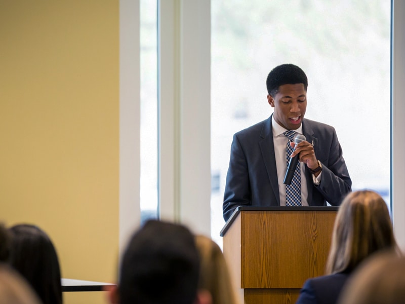 A student in a suit speaks confidently into a microphone at a podium during a leadership event.