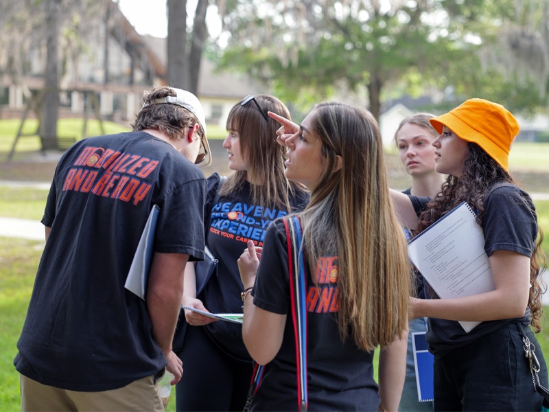 A group of students collaborate outdoors, discussing notes and pointing directions during a team activity.