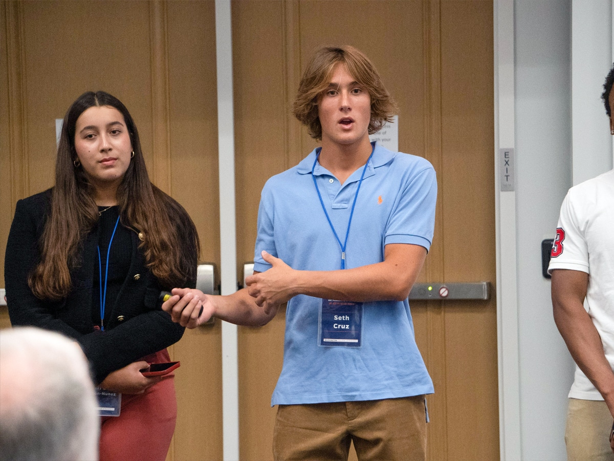 A student gestures with his hands while presenting during a classroom session, as another student stands beside him listening.