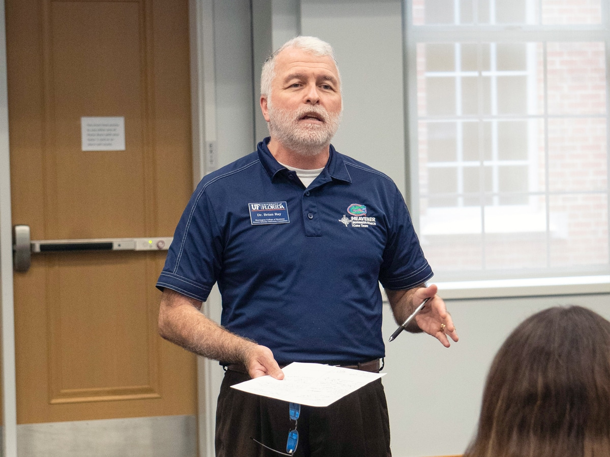 A faculty member speaks to students in a classroom, holding a pen and paper while emphasizing a point during a Management Communication Center course.
