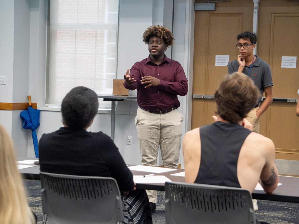 A student stands and speaks confidently with animated gestures during a classroom presentation, while peers and instructors observe from seated positions.