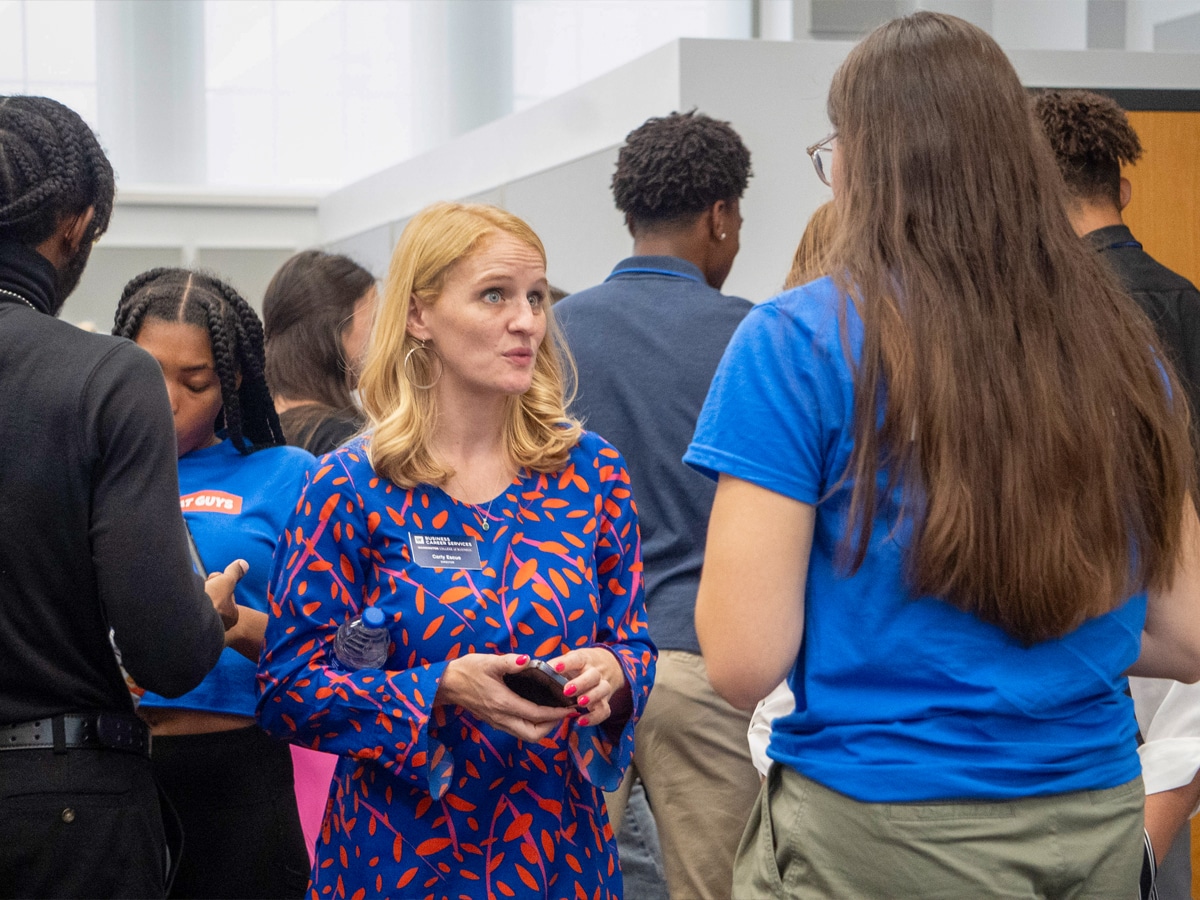 A faculty member in a patterned dress speaks with a student in a crowded classroom setting during a Management Communication Center workshop.