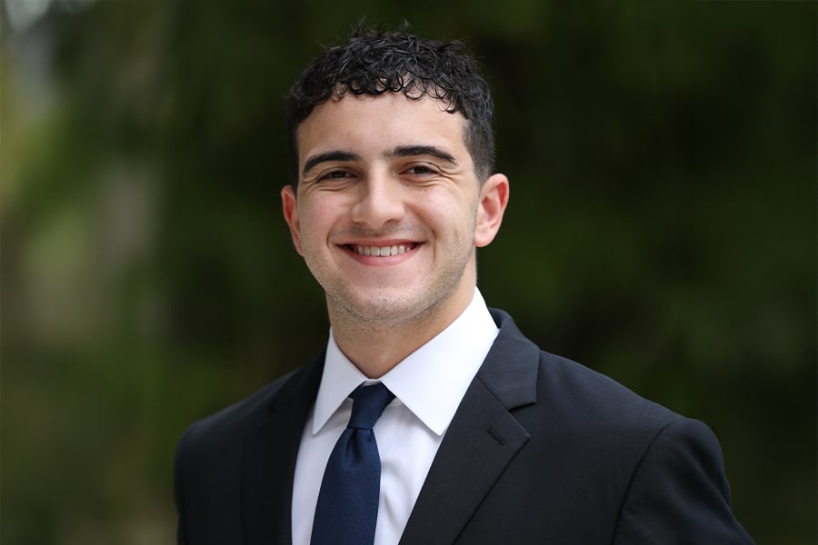 A young man in a black suit, white shirt, and dark blue tie smiles warmly while standing outdoors against a softly blurred green background.