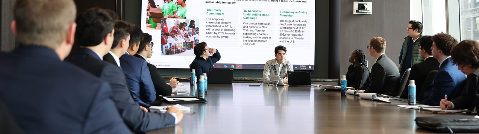 A group of business people gathered around a conference table for a meeting, with a large screen display visible in the background.