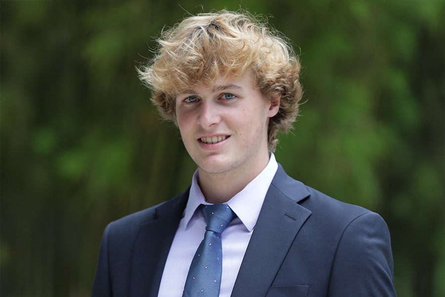 A young man with curly blond hair wearing a dark suit, lavender shirt, and patterned tie smiles slightly while standing outdoors in front of a blurred green background.