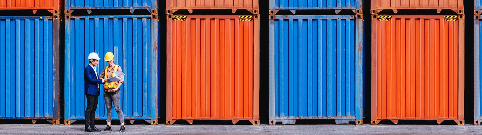 Two supply chain professionals in hard hats stand in front of blue and orange shipping containers