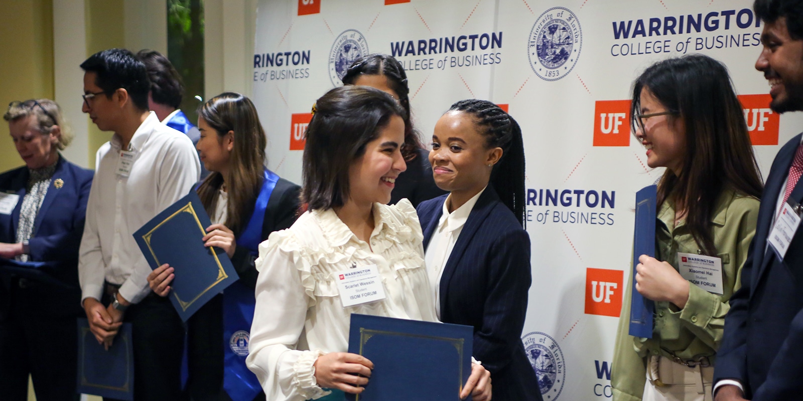 After completing their practicum project, students from UF Warrington's MS in ISOM - Supply Chain Management program hold certificates and smile at each other while assembling themselves in front of a wall with the school's logo for a photo