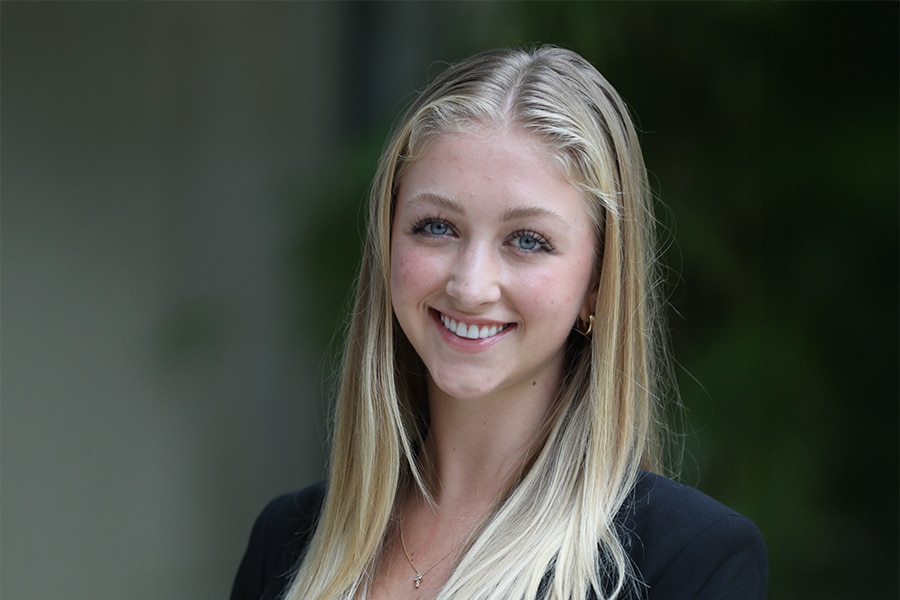 A young woman with long blonde hair, wearing a black blazer and gold earrings, smiles confidently in an outdoor setting with a soft green background.
