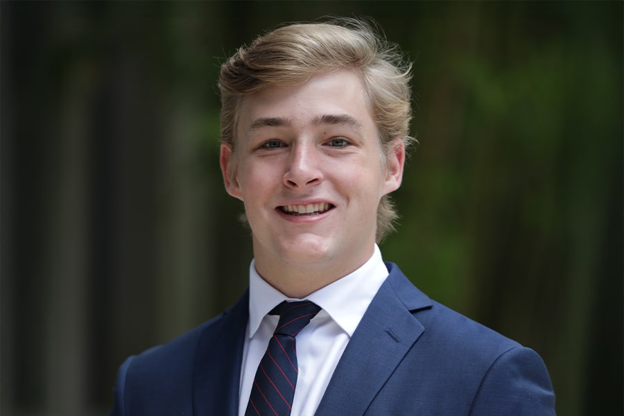 A professionally dressed young man in a navy blue suit and striped tie smiles confidently in an outdoor setting with soft-focus greenery in the background.