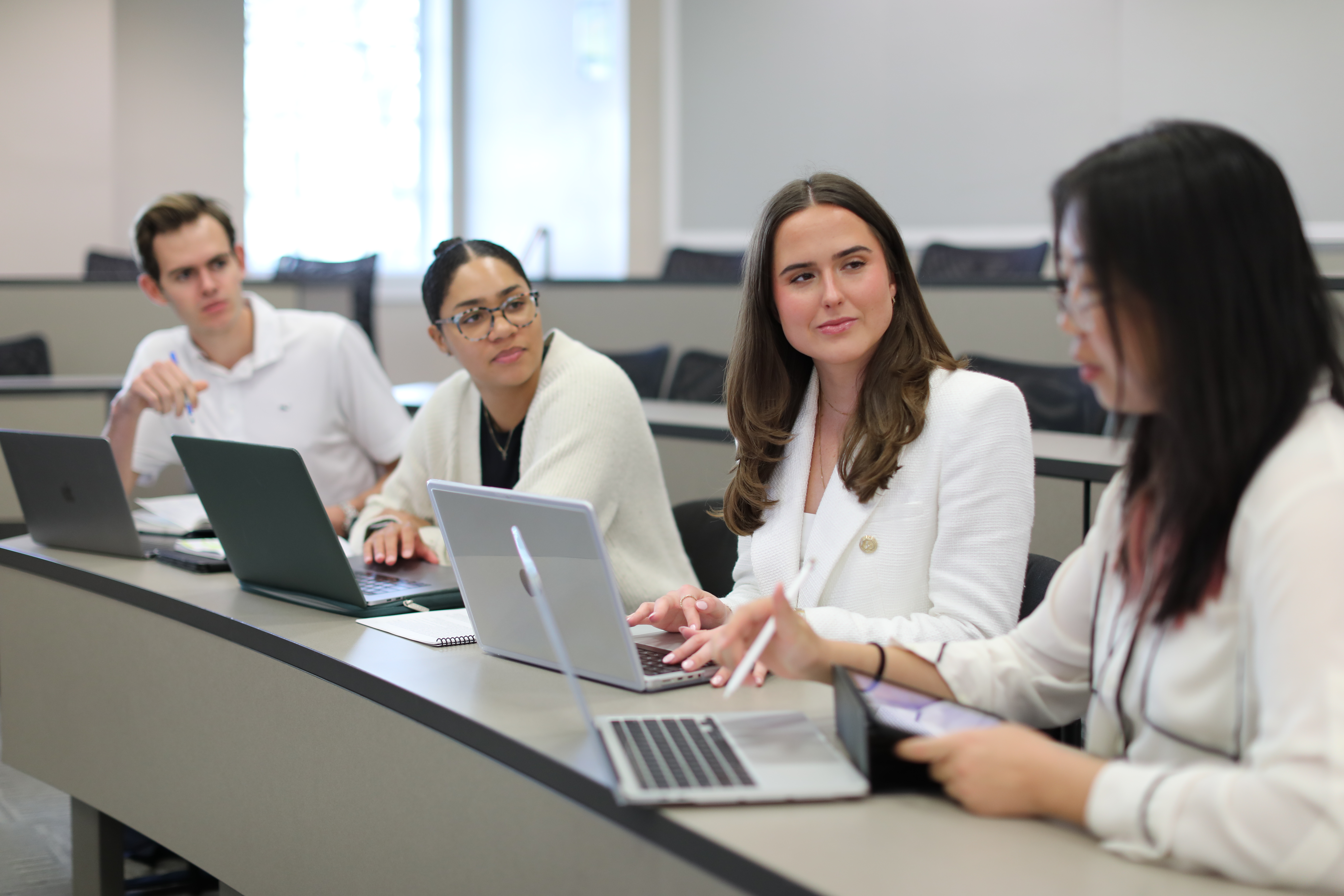Four students sit at a curved desk with laptops, engaged in a collaborative discussion involving digital tools.