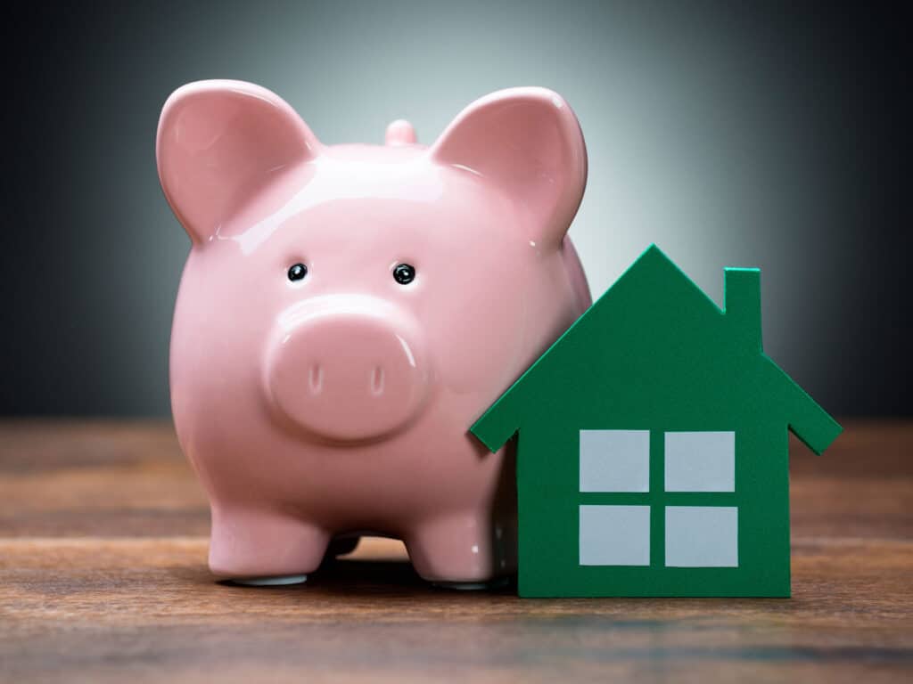 A tiny piggy bank next to a 2D green house shape