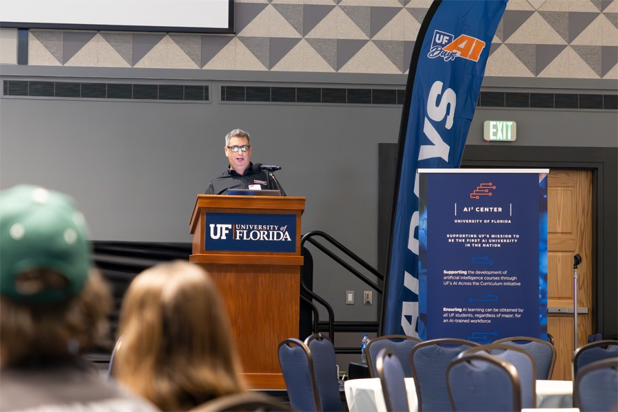 A speaker in glasses addresses a room from a wooden podium with the University of Florida logo, as a group of people watch from the audience. Banners for UF AI Days and the AI Center stand nearby.