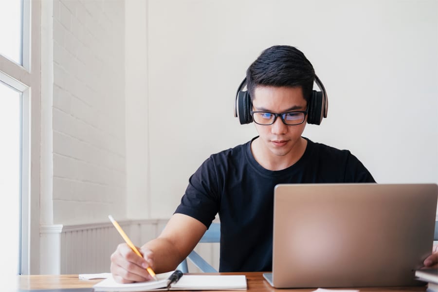 A student wearing headphones and glasses works on a laptop while taking notes with a pencil in a notebook at a sunlit desk.