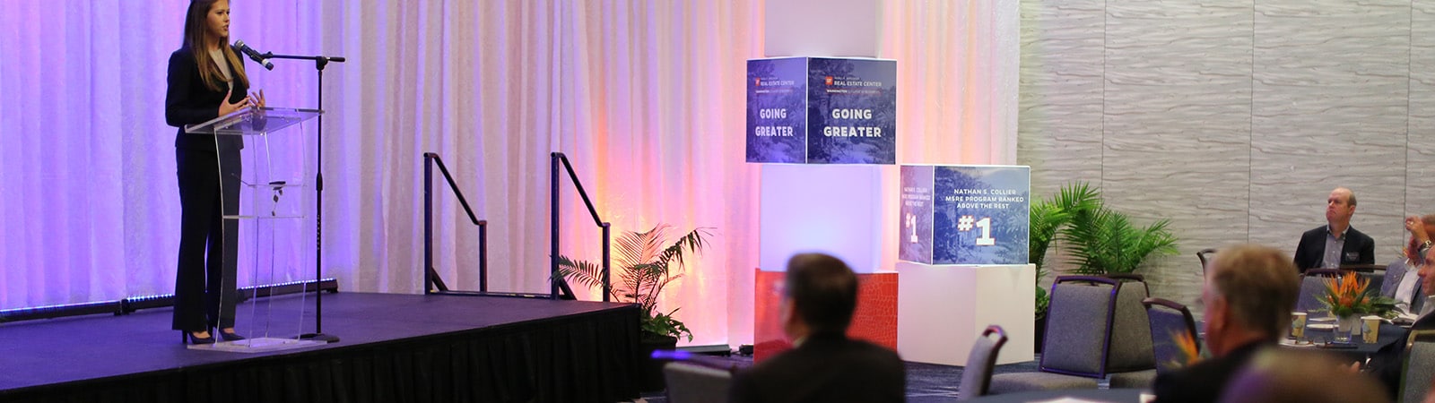 Woman speaking on stage at a conference with an audience of people sitting at tables in front of the stage.