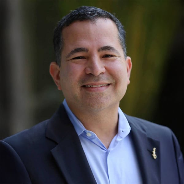 Portrait of Angel Arroyo, a smiling man with short black hair, wearing a dark blazer over a light blue shirt.