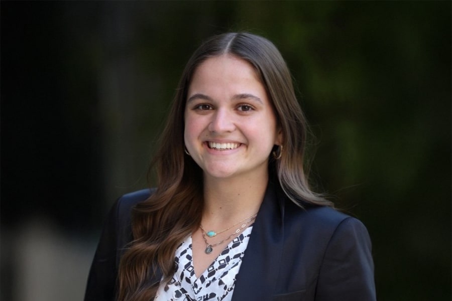 Aubrey Harris, President of Business Ethics Ambassadors, smiles for a professional headshot, wearing a dark blazer over a patterned blouse, with long brown hair and layered necklaces.