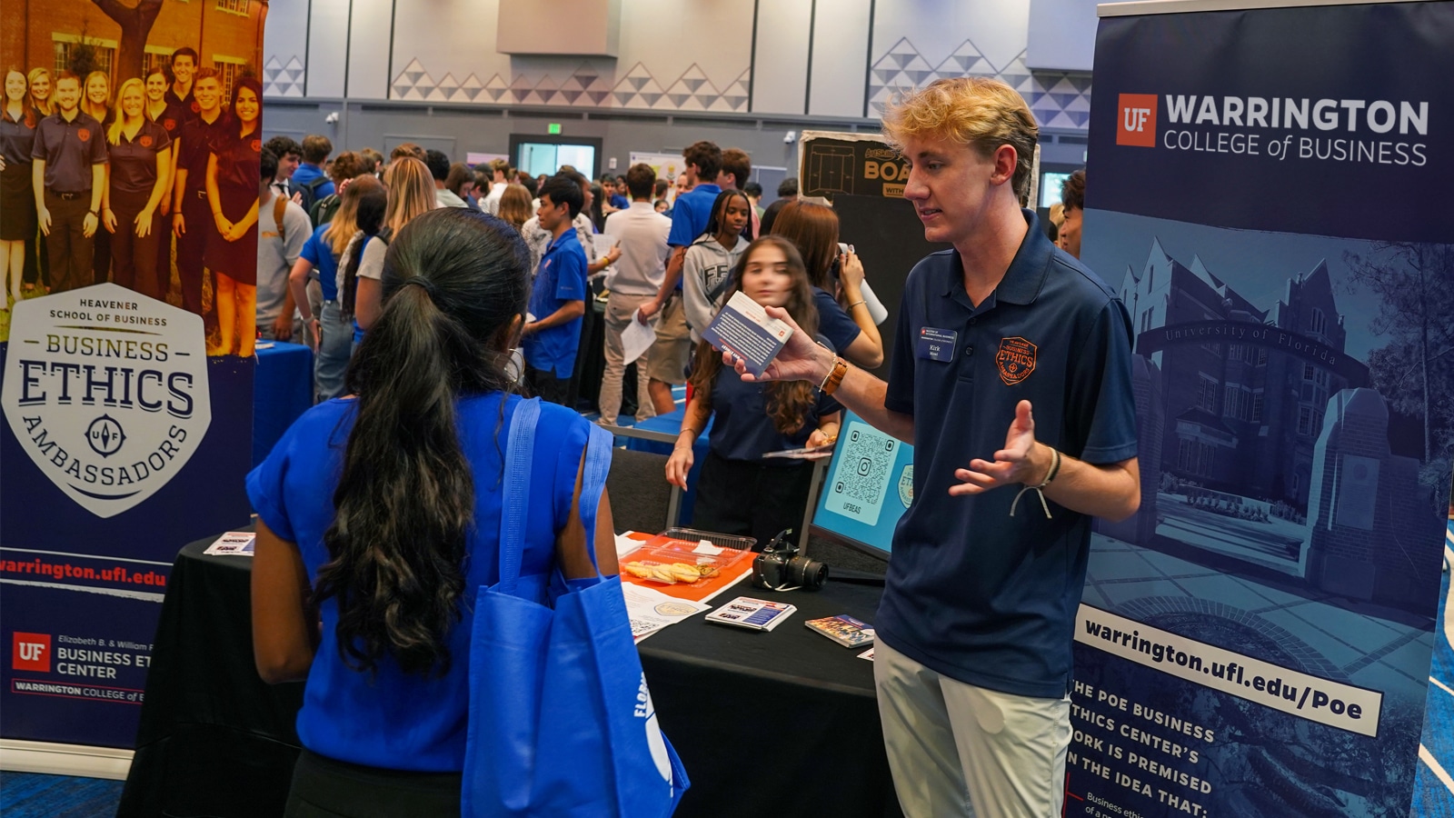 A student in a navy Business Ethics Ambassadors polo shirt talks with an attendee at a crowded campus event, with informational banners and materials for the Business Ethics Center on display.