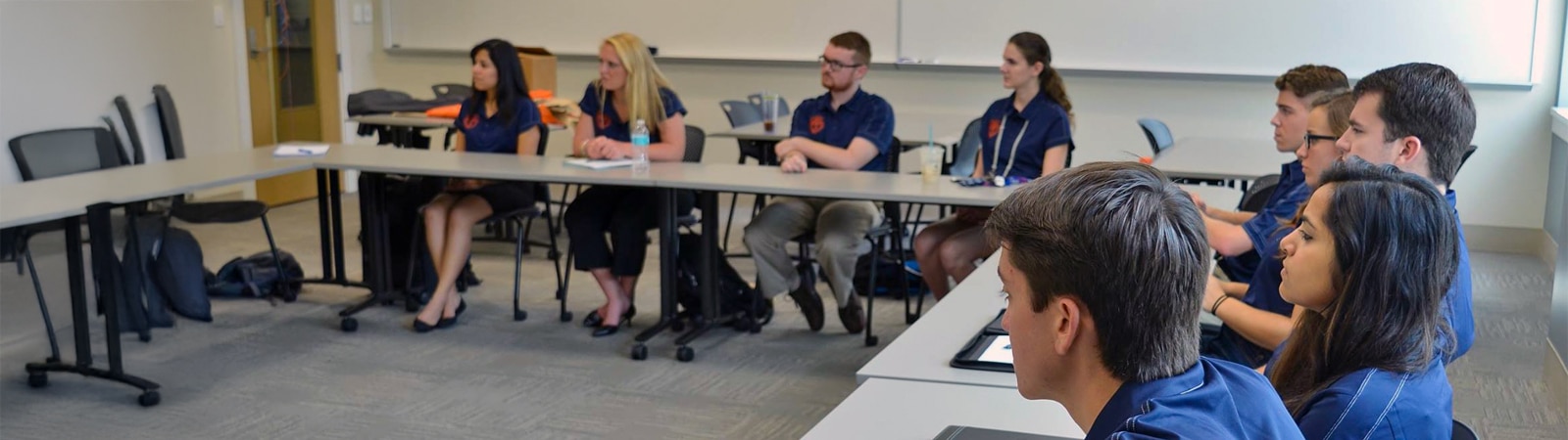 A group of students in matching shirts sit around conference tables in a classroom, attentively listening to a discussion about business ethics.