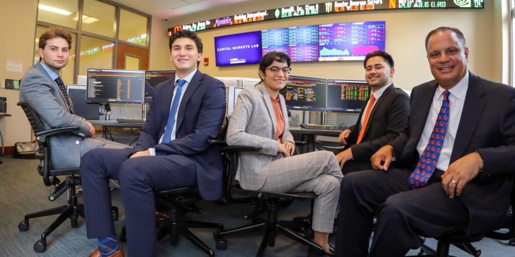 Wearing suits, UF Warrington students and faculty participating in the Caimanes Student Managed Equity Hedge Fund pose in front of computer monitors in a trading simulation space