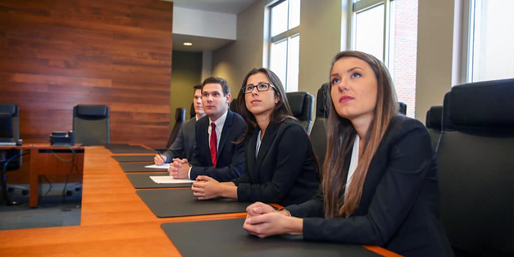 Wearing suits, students from UF Warrington's BSBA in Finance program sit around a boardroom table while looking upward during a presentation