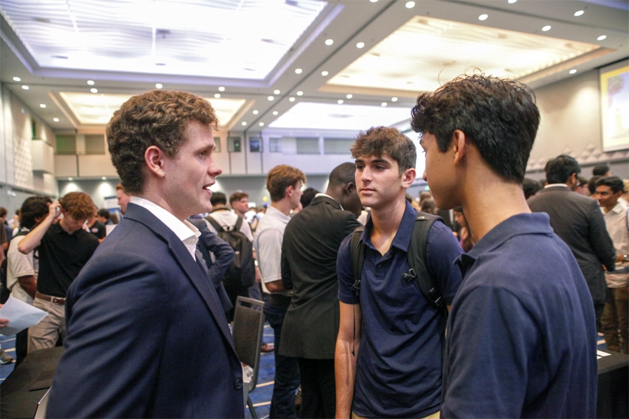 A young professional student in a navy suit speaks to two younger students in a crowded event space, surrounded by peers networking and socializing.