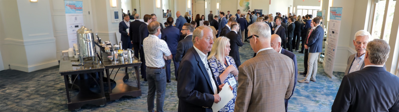 A busy networking event with groups of business professionals engaged in conversation in a spacious, well-lit conference room with large windows.