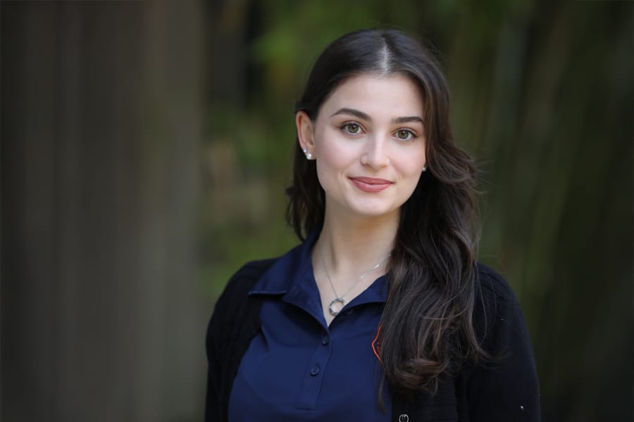 Bianca Barroso, Director of Merchandise for Business Ethics Ambassadors, poses outdoors in a professional headshot, wearing a navy blue polo and black sweater.