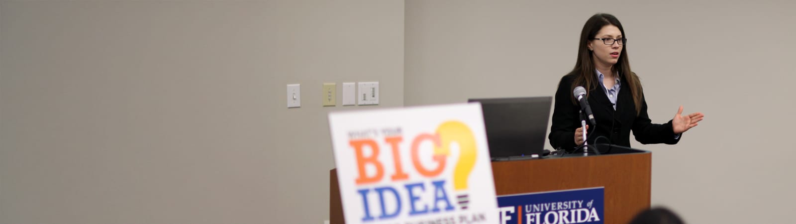 Young woman in professional attire presenting at a podium with a laptop, speaking to an audience. In the foreground, a sign reads "What's your BIG IDEA?".