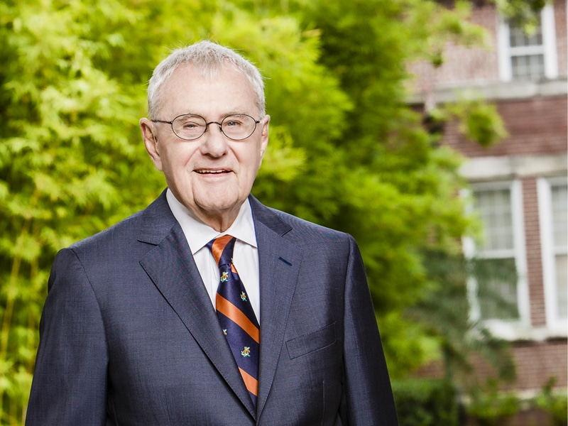 Bill Hough in a dark suit and colorful tie, stands outdoors in front of a green background with a brick building partially visible.