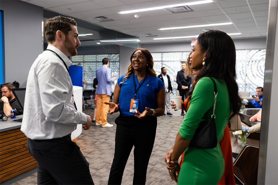 A group of diverse professionals are seen engaging in animated conversation in a modern, collaborative office space with bright lighting and large windows.