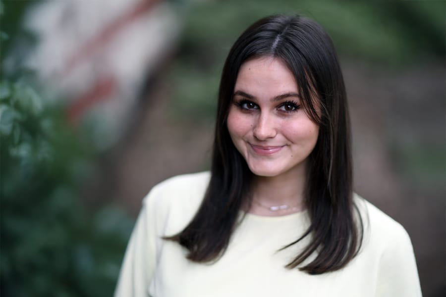 Portrait of Bri Urukalo, a young woman with dark hair wearing a light-colored top, smiling softly outdoors with a blurred natural background.