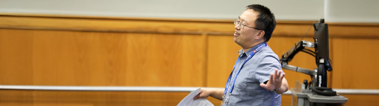 A professor wearing glasses and a Warrington College of Business lanyard gestures expressively while teaching in a lecture hall, holding papers in one hand.