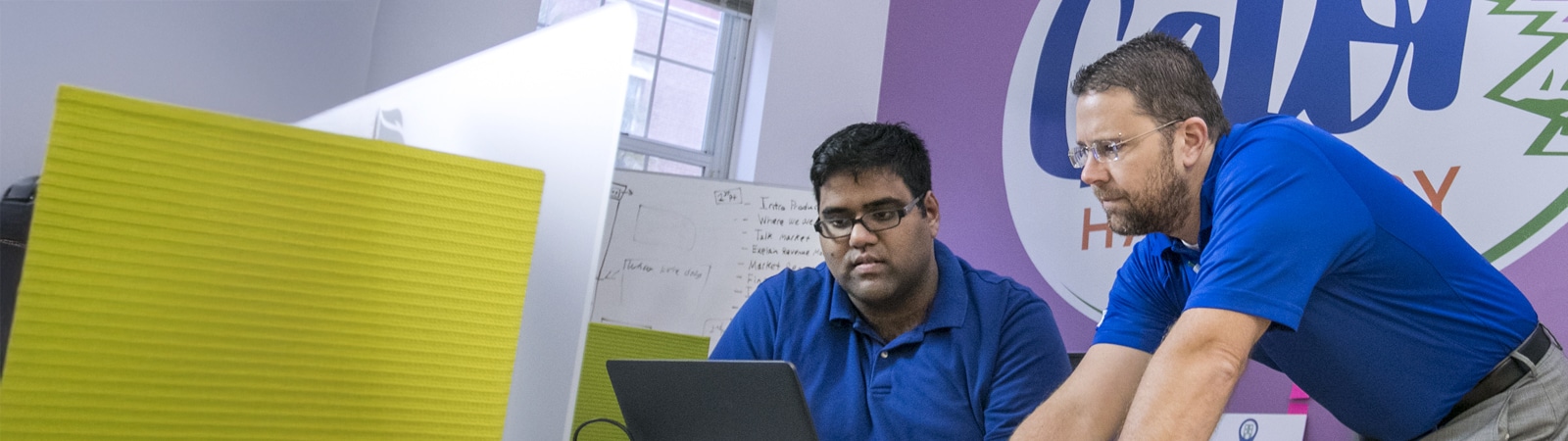 Two men collaborate in a brightly colored workspace. One is seated and working on a laptop while the other leans in to discuss ideas. They’re wearing blue shirts and focused on the task at hand, with a vibrant “Gator Hatchery” sign in the background.