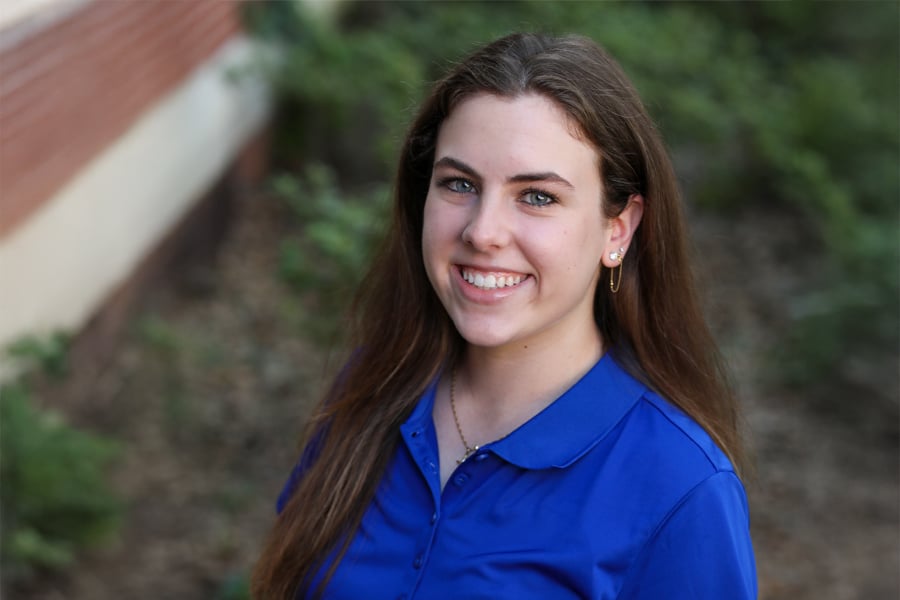 Portrait of Carly Rutigliano, a smiling young woman with long brown hair wearing a royal blue polo shirt, standing outside near greenery.