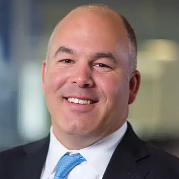 Headshot of Chris McGibbon, a middle-aged man with a shaved head and light skin, smiling and wearing a black suit jacket, white shirt, and light blue tie. The background is softly blurred and indoors.