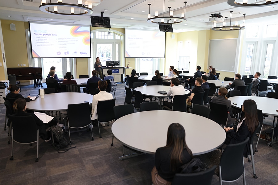 A woman speaking to a class of professionally dressed students. 