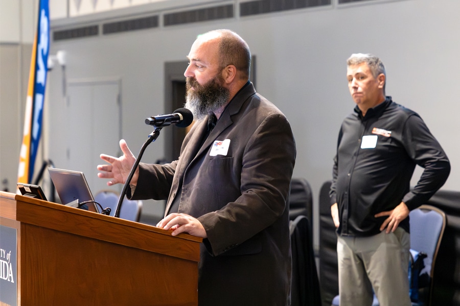A bearded speaker in a suit presents at a podium with a microphone, while another man in a black shirt stands attentively in the background at a business conference.