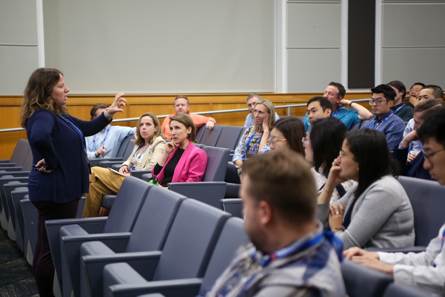 A woman stands at the front of an auditorium, engaging a diverse group of faculty and professionals in lively discussion, with audience members listening and asking questions.