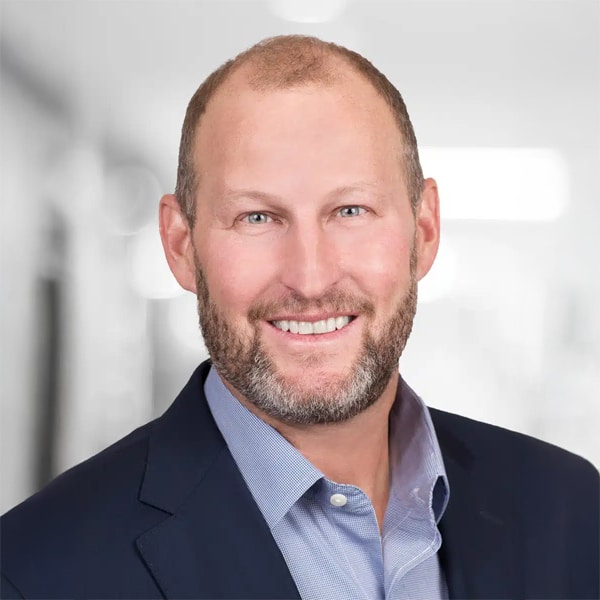 Portrait of Dirk Aulabaugh, a smiling man with short hair and beard, wearing a dark blazer over a light blue shirt, against a softly blurred background.