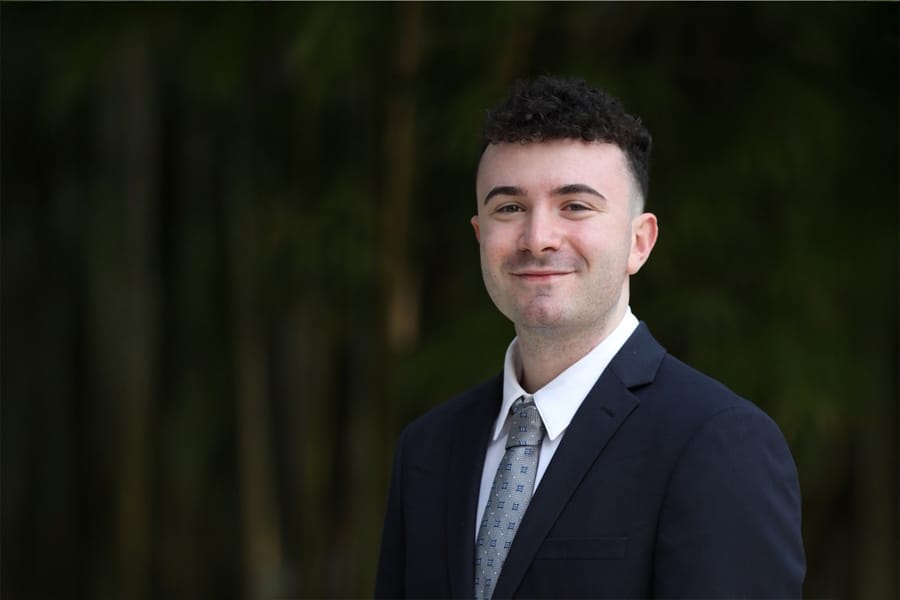 A young man with short curly hair, wearing a dark suit jacket, white shirt, and patterned tie, smiles in a professional outdoor portrait.