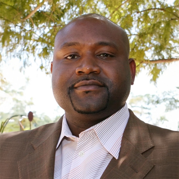Portrait of Eddy Benoit, Jr., a man with a closely shaved head and neatly trimmed facial hair, wearing a brown suit jacket over a white striped shirt, standing outdoors with trees softly blurred in the background.